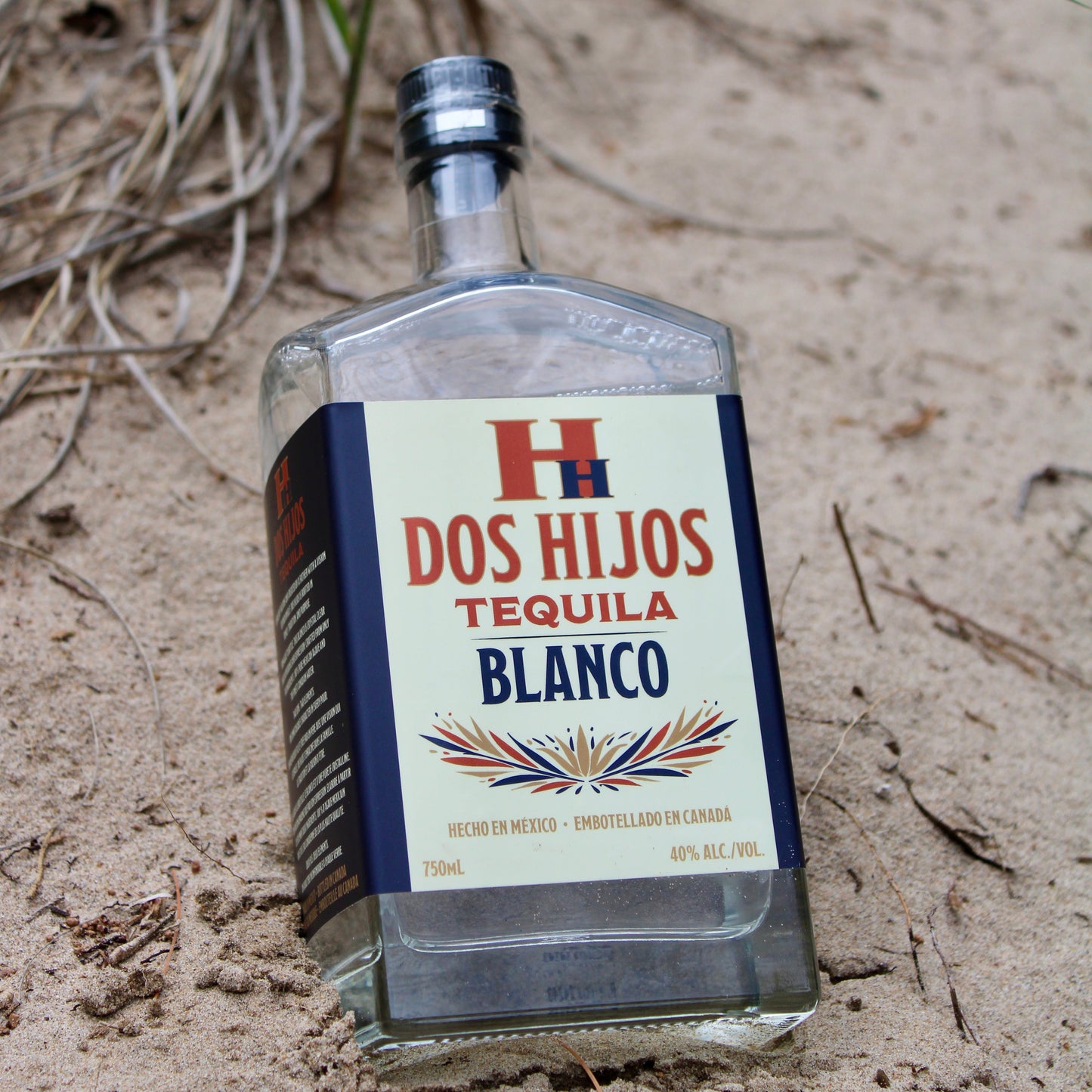 Dos Hijos Tequila Blanco bottle on sandy ground.